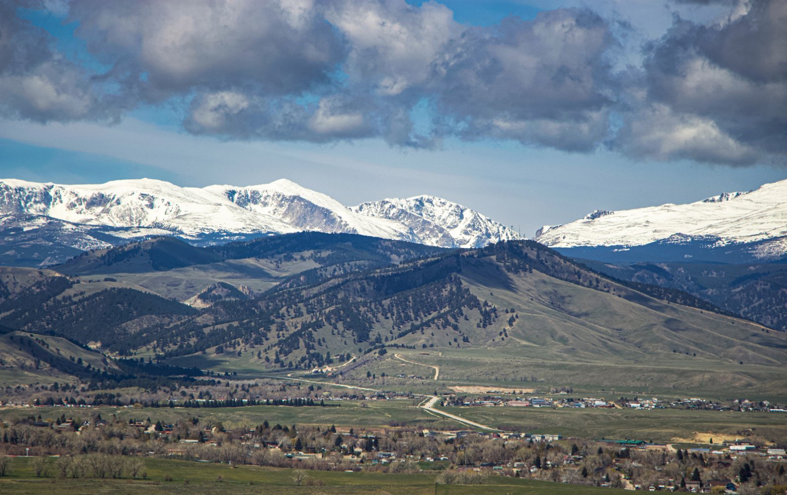 Scenic valley landscape with snow-capped mountains in the distance, green fields, scattered homes, and cloudy sky overhead